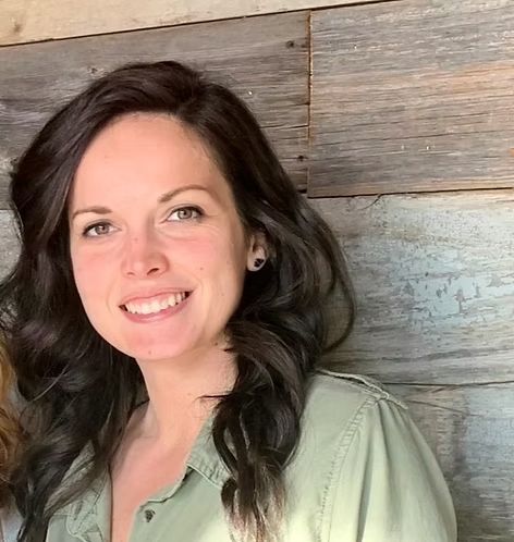 A woman in a green shirt is smiling in front of a wooden wall.