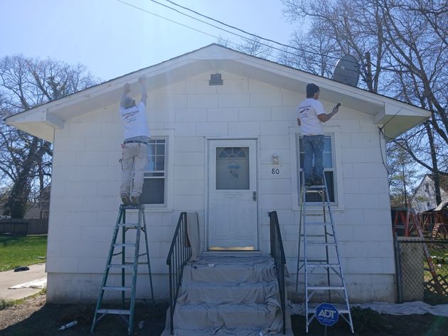 Two workers on ladders painting the front of a small white house.