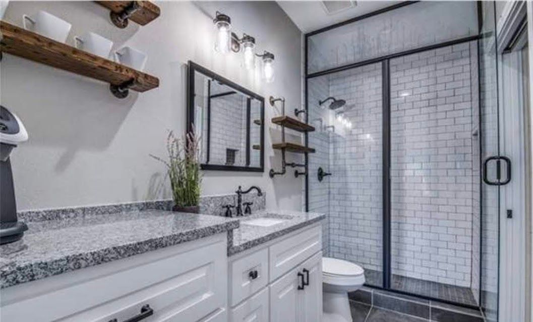Modern bathroom with white vanity, granite countertop, black-framed mirror, shelves, toilet, and glass shower.
