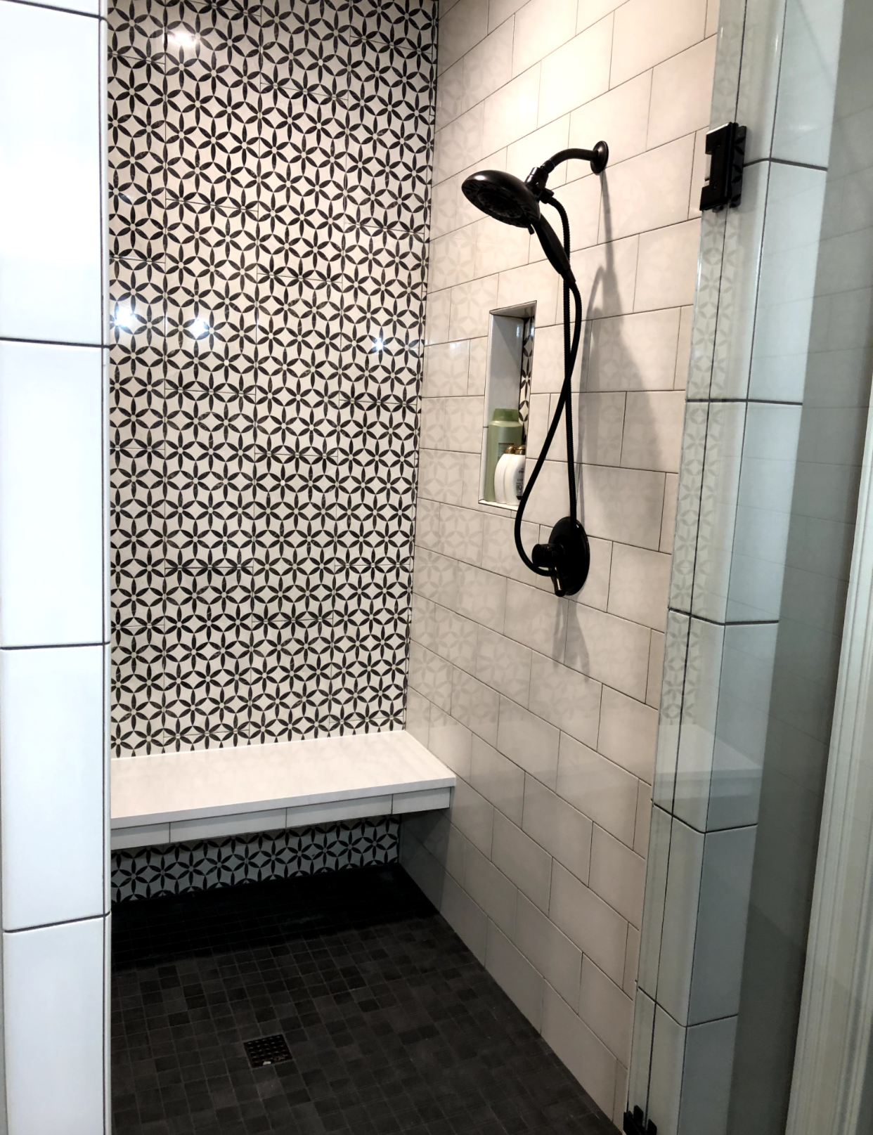 Small tiled shower with black fixtures, bench, patterned accent wall, and dark floor drain.