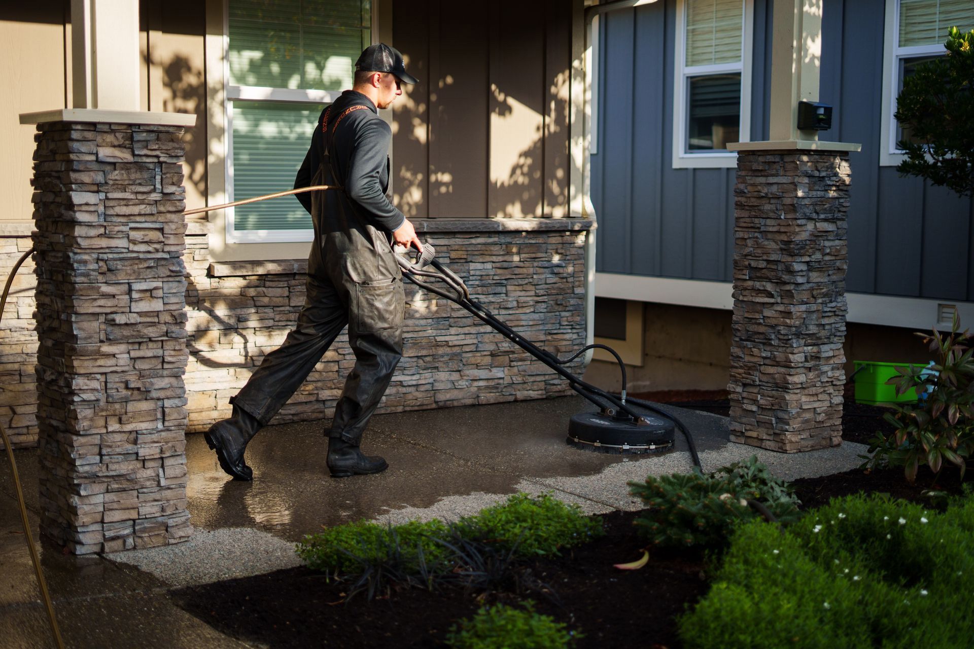 Man pressure washing a concrete patio next to a house with stone pillars and landscaping.