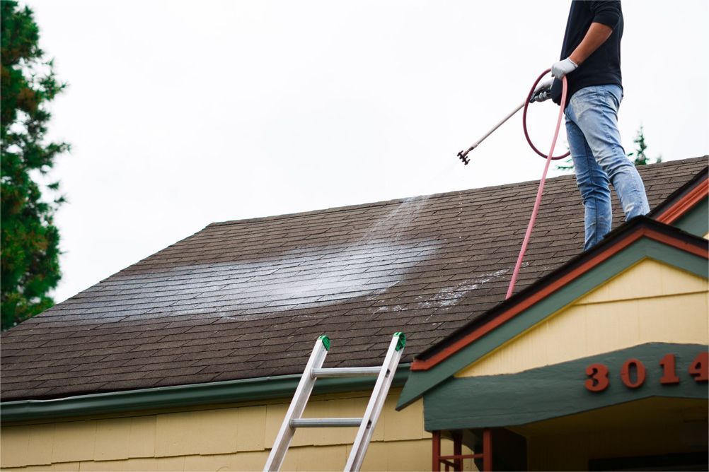 Person on roof washing it with a hose, using ladder for access; beige house, cloudy sky.