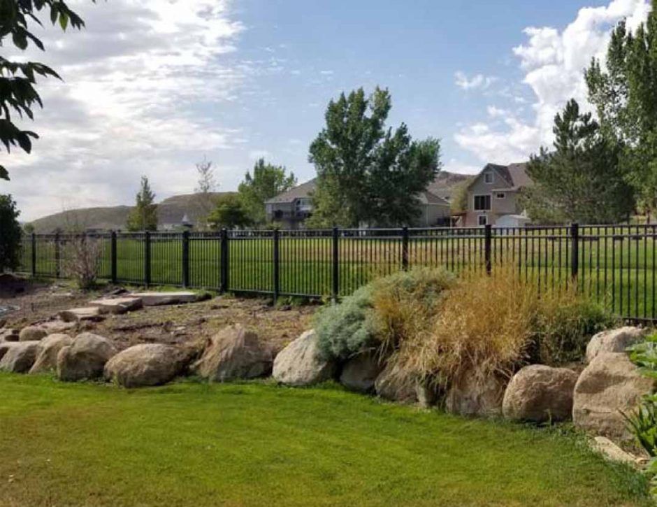 A black fence surrounds a lush green yard with rocks and trees.