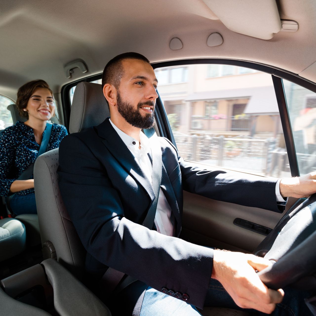 A man in a suit is driving a car with a woman in the back seat.