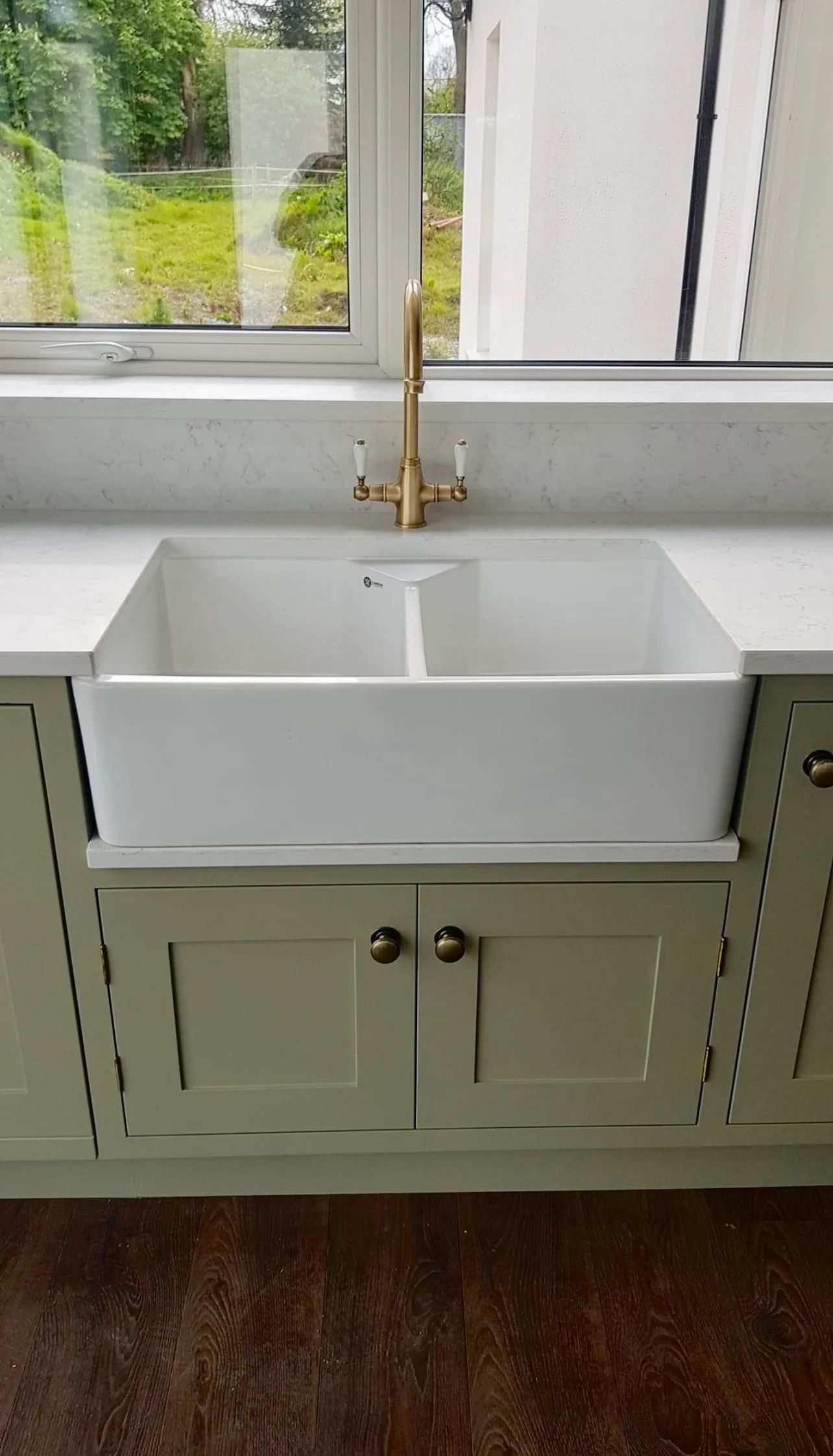White farmhouse sink with gold faucet, sage green cabinets, and a window.