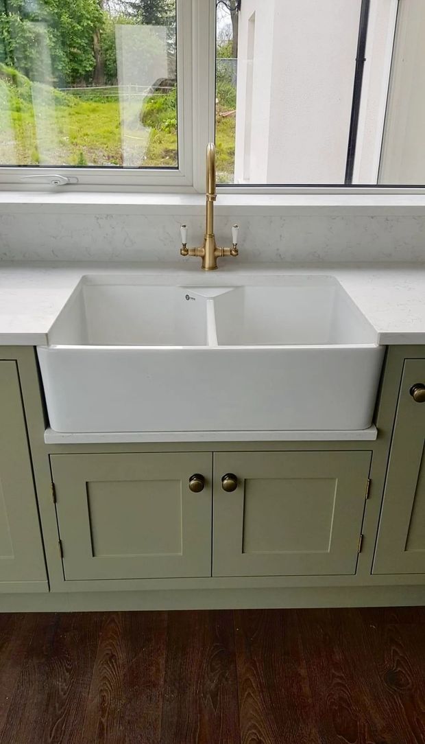 White farmhouse sink with gold faucet, sage green cabinets, and a window.