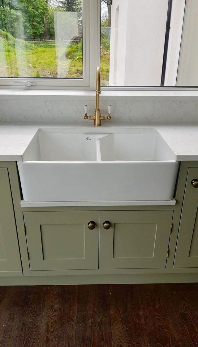 White farmhouse sink with gold faucet, sage green cabinets, and a window.