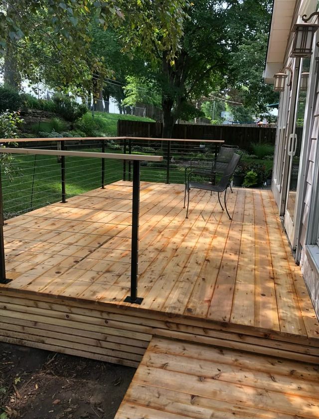 Wooden deck with black metal railing and cable infill, overlooking a green yard and trees.