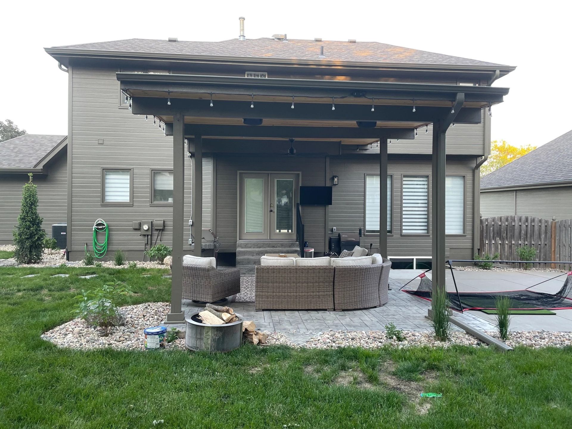 Backyard patio with a pergola, seating, and a house in the background. Green grass and landscaping are in the foreground.