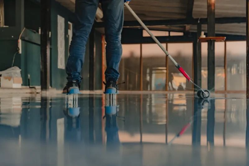 Person applying blue epoxy to a floor with a roller. Outdoors, sunset reflecting on the surface.