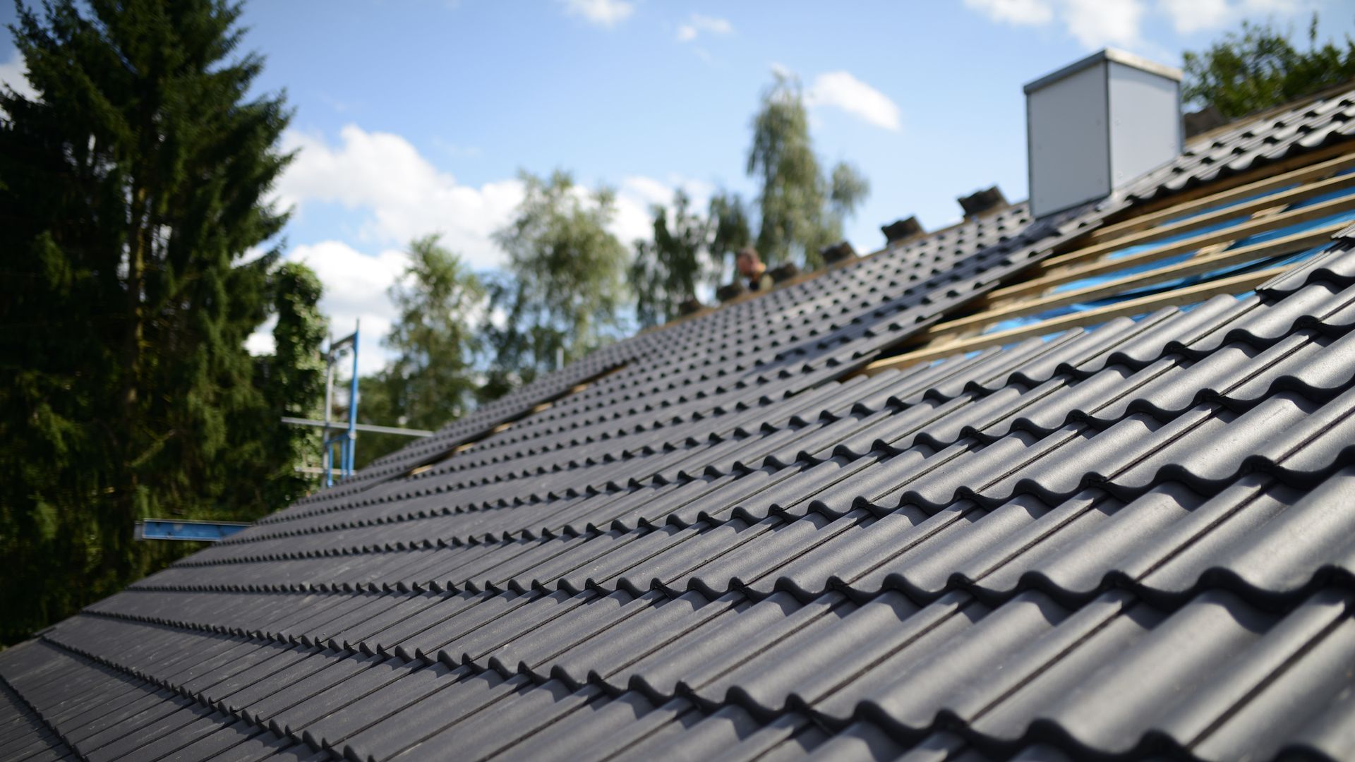 Close-up of a dark gray tiled roof with a chimney under a blue sky, trees in the background
