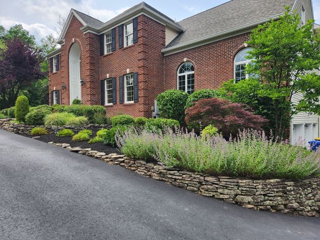 A two-story red brick house with a stone retaining wall, paved driveway, and varied garden landscaping.
