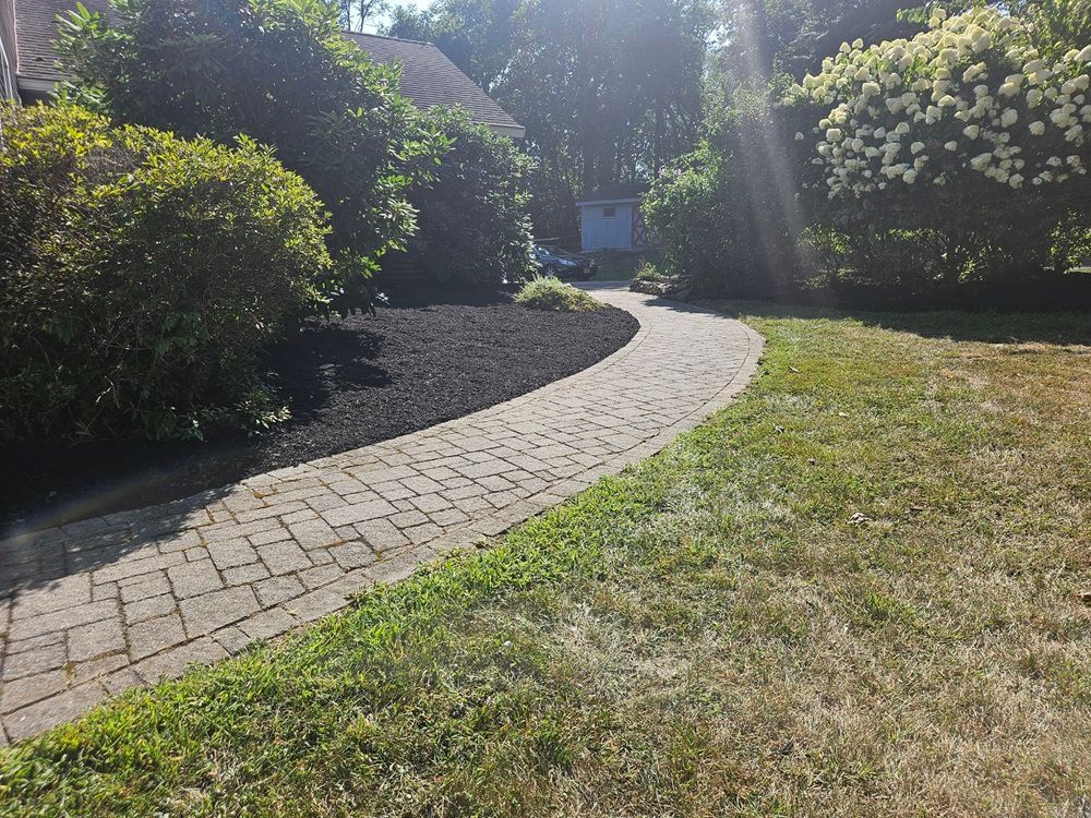 A stone paver walkway curves through a front yard, bordered by dark mulch, green bushes, and a patch of grass.