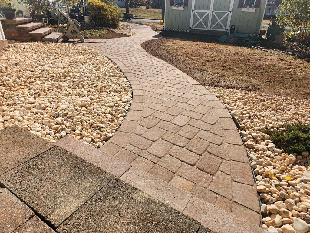 A tan interlocking paver walkway curves through a yard with light-colored landscaping stones and a shed in the background.