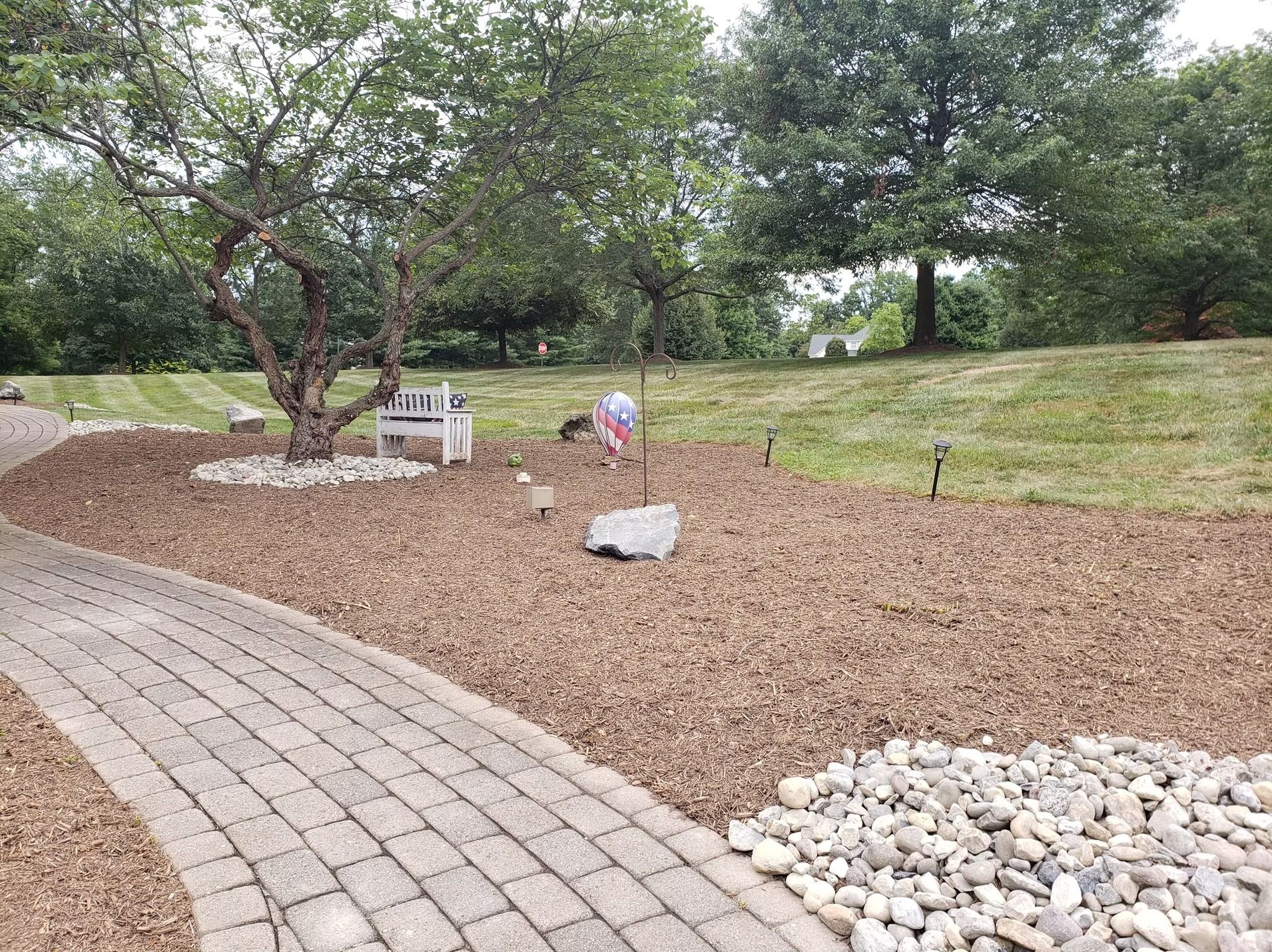 A paved walkway curves through a mulched landscape with a tree, a decorative bench, a small boulder, and scattered lights.