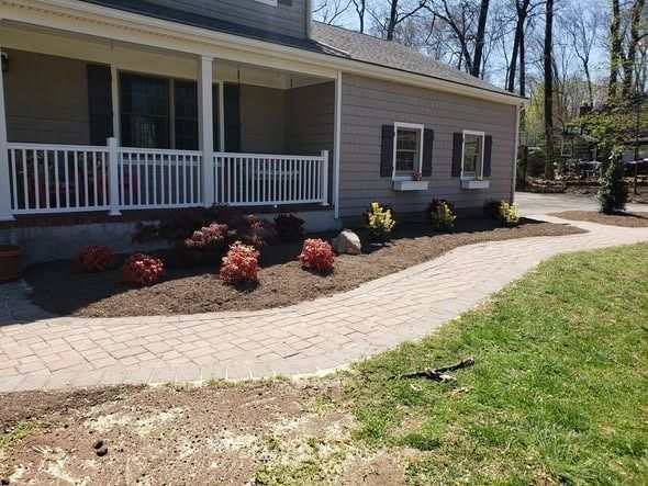 A paved stone walkway curves toward the porch of a gray house with dark shutters and freshly mulched garden beds.