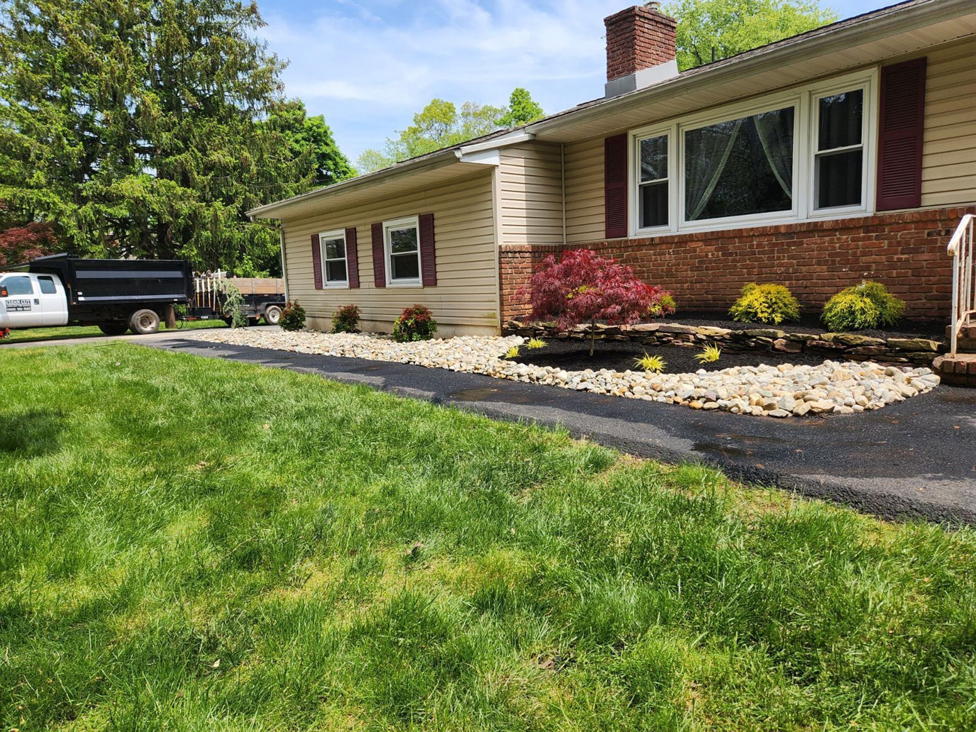 A one-story house with tan siding, a brick facade, and a red maple bush in a landscaped rock garden bed by the driveway.
