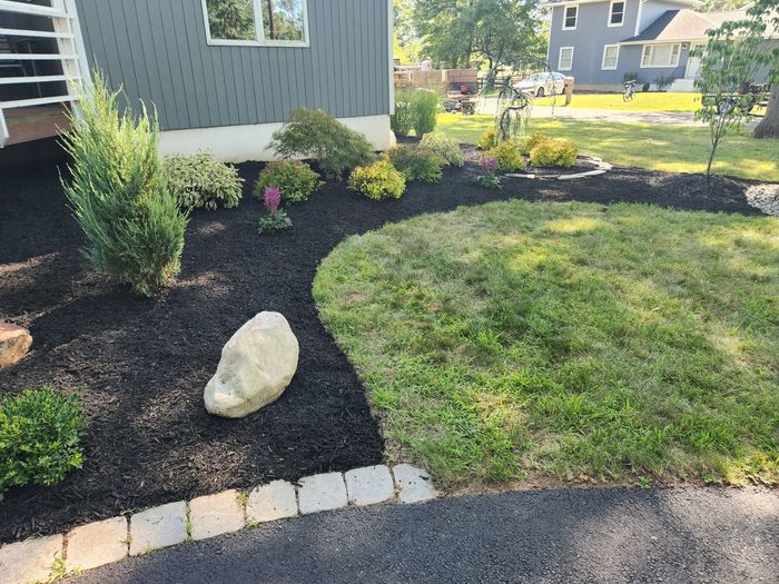 A light-colored house with cedar shingles, a porch, and a driveway bordering a garden bed filled with black mulch.