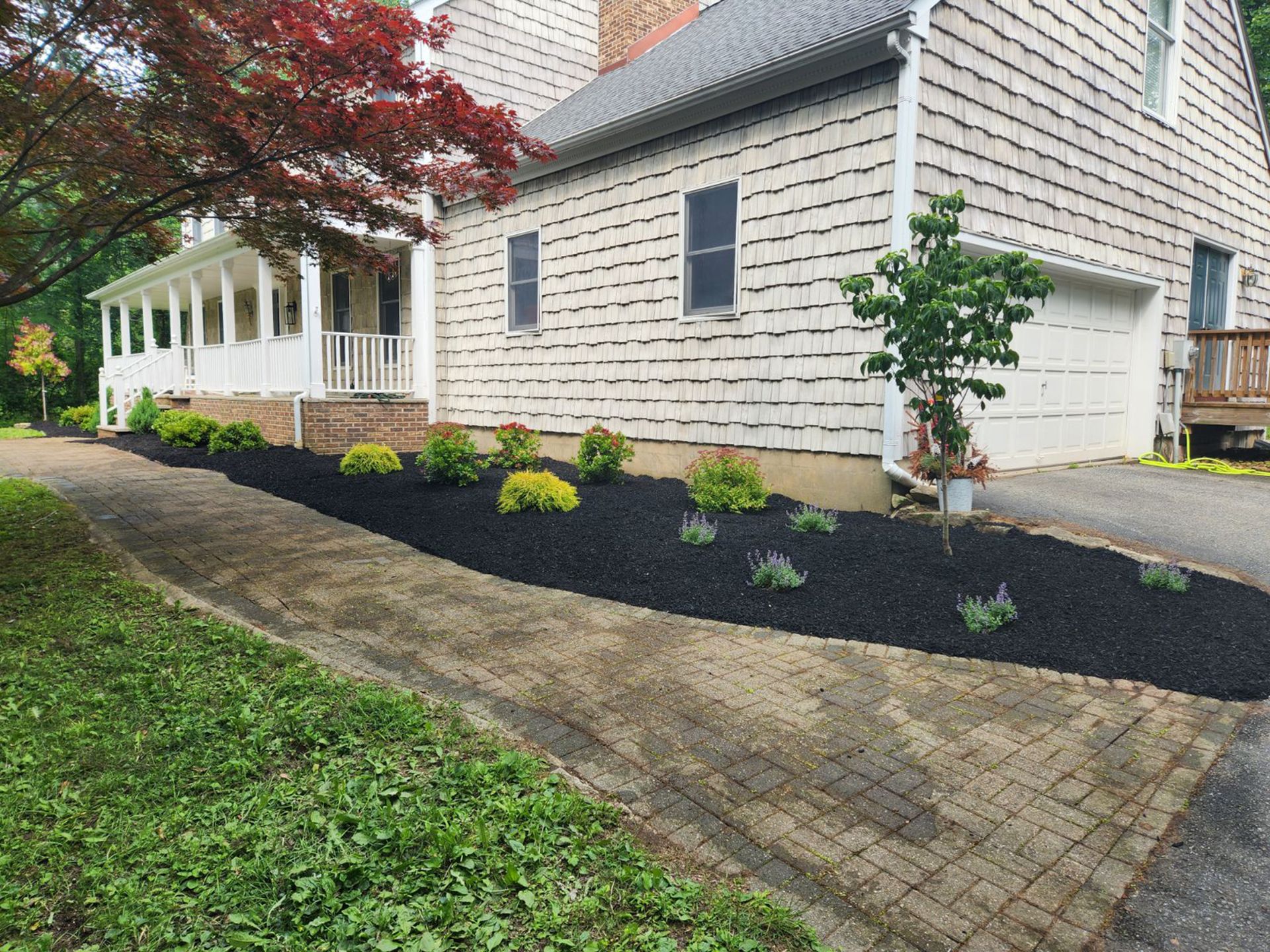 A light-colored house with cedar shingles, a porch, and a driveway bordering a garden bed filled with black mulch.