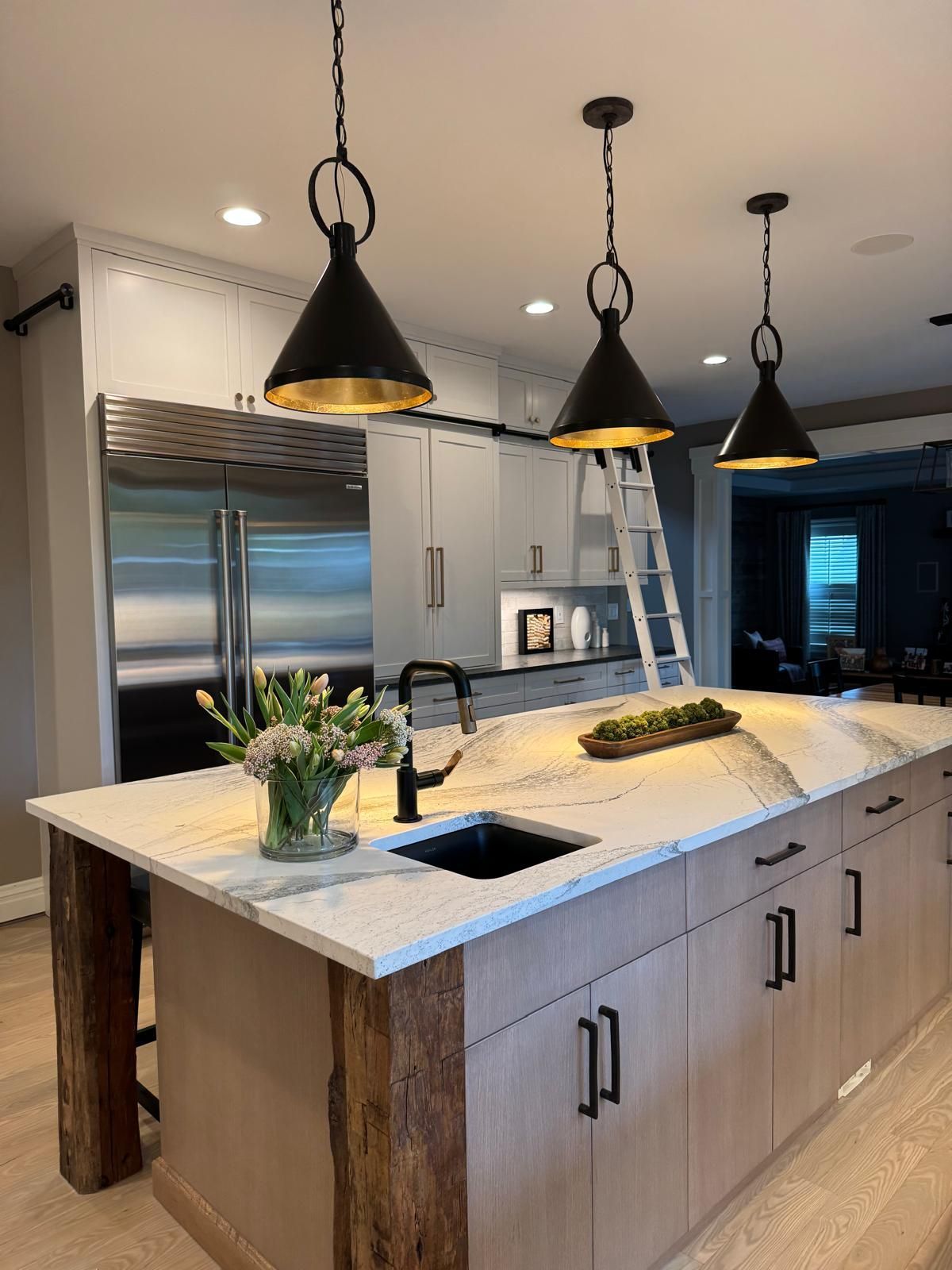 Modern kitchen with large island, pendant lights, stainless fridge, and a ladder by the cabinets.