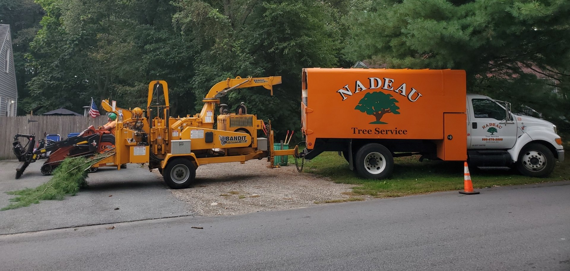 Orange and white utility truck with wood chipper parked by roadside, worker clearing brush and cones nearby