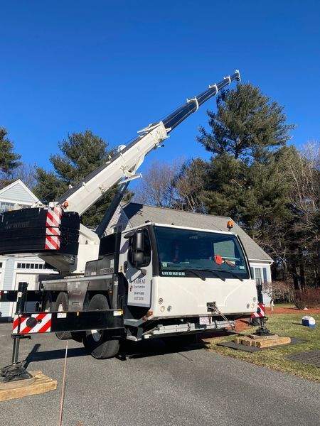 A white mobile crane with an extended boom parked on a residential driveway, stabilized by outriggers on wooden pads.