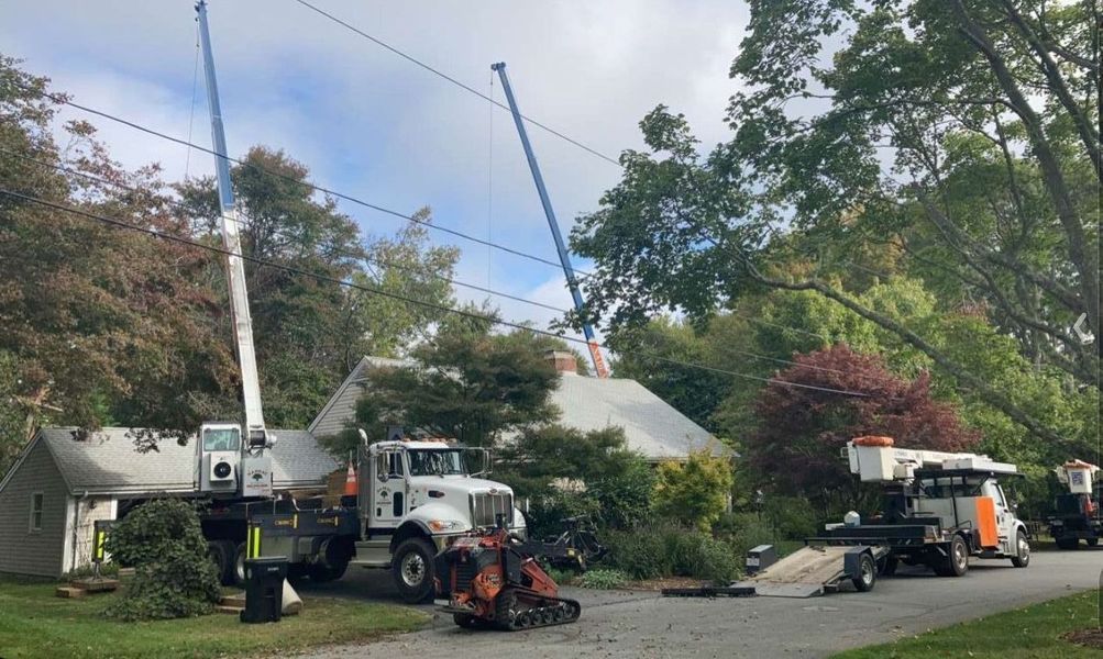 Two utility trucks with tall cranes and a wood chipper parked in front of a residential house with trees.
