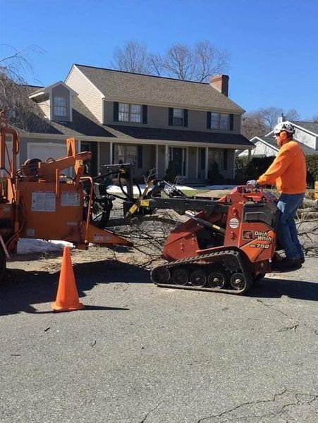 A worker in an orange shirt operates a tracked wood chipper on a suburban street in front of a house.