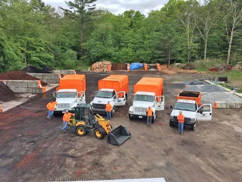 A team of workers in high-visibility orange shirts stands with three orange dump trucks and a loader in a landscape yard.