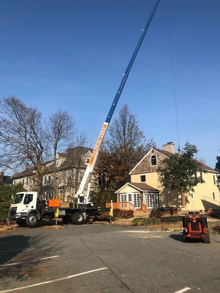 A large crane truck is parked on a paved lot, with its boom extended high toward a multi-story house on a clear, sunny day.