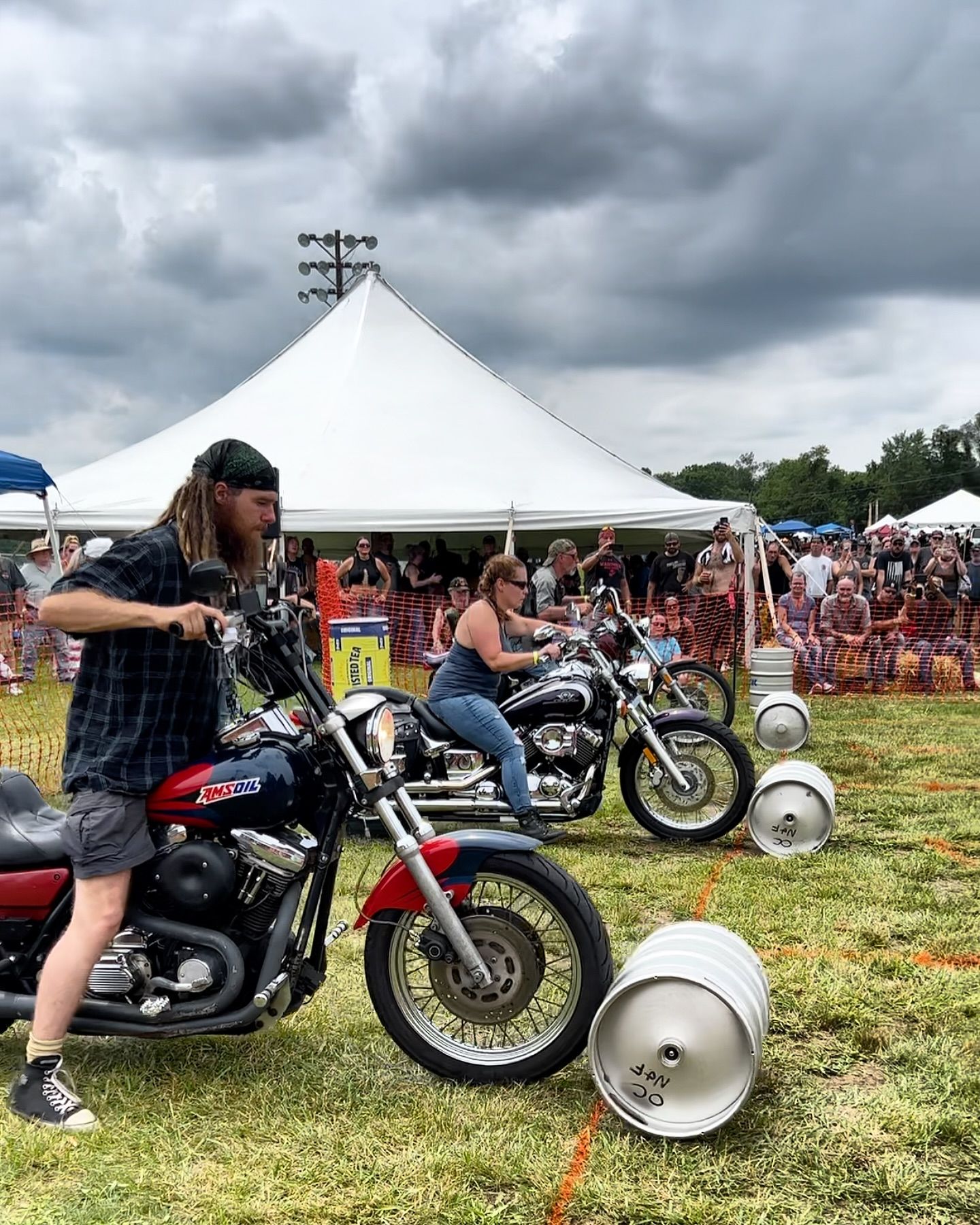 Two people on motorcycles maneuvering around beer kegs in a field, with a crowd and white tent in the background.