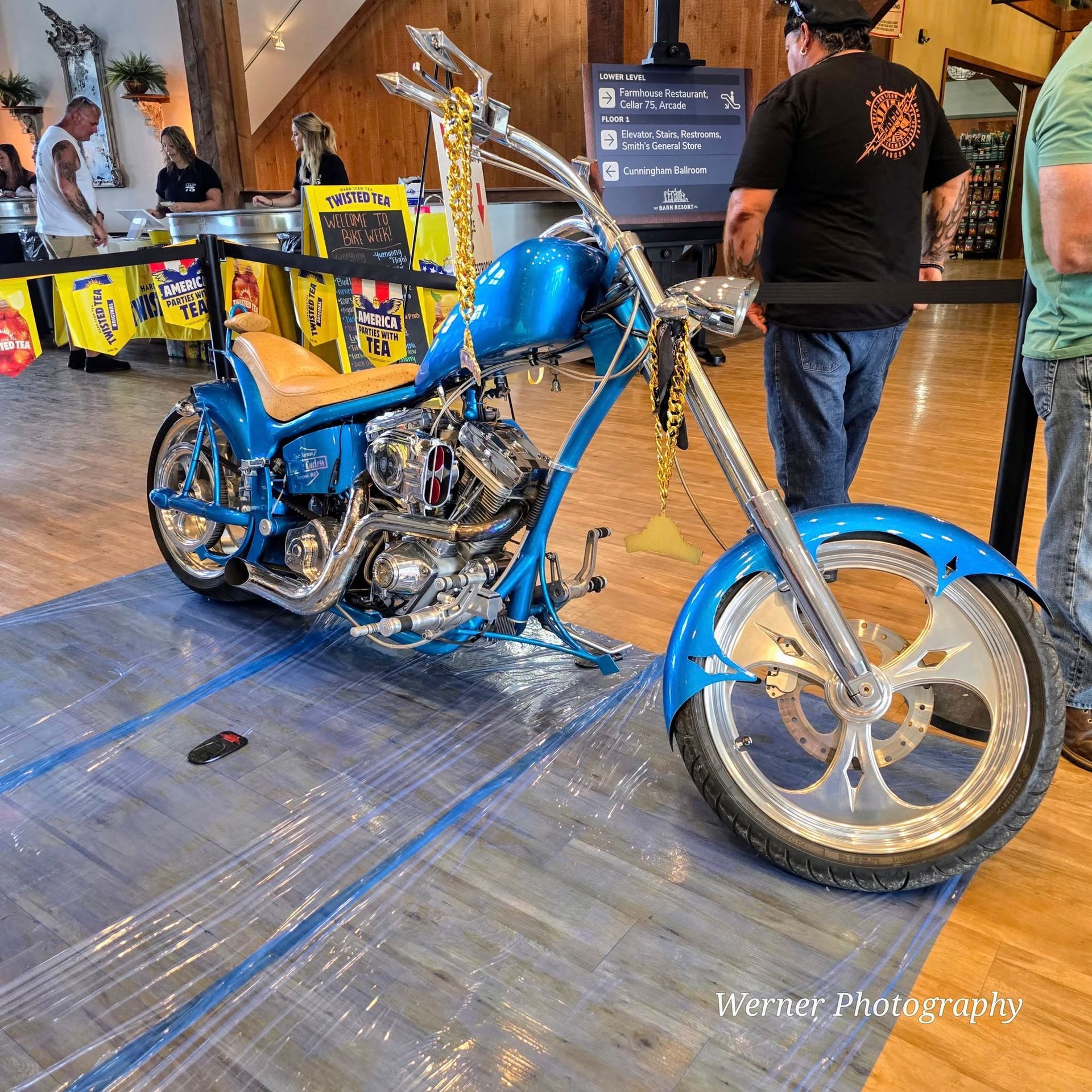 Blue custom motorcycle on display at an event with people, a light brown building, and a clear plastic covering.