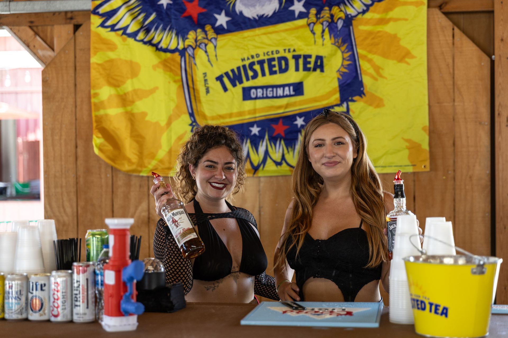 Two women behind a bar serving Twisted Tea. One holds a bottle, the other smiles. A Twisted Tea flag is behind them.