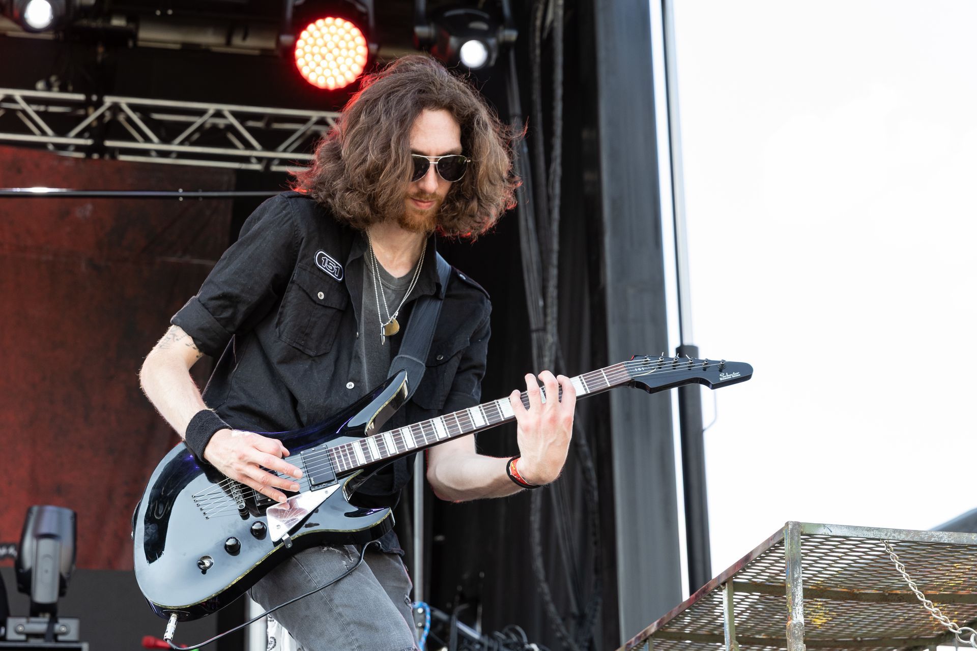 Guitarist with long hair, sunglasses, and black shirt playing electric guitar on outdoor stage.