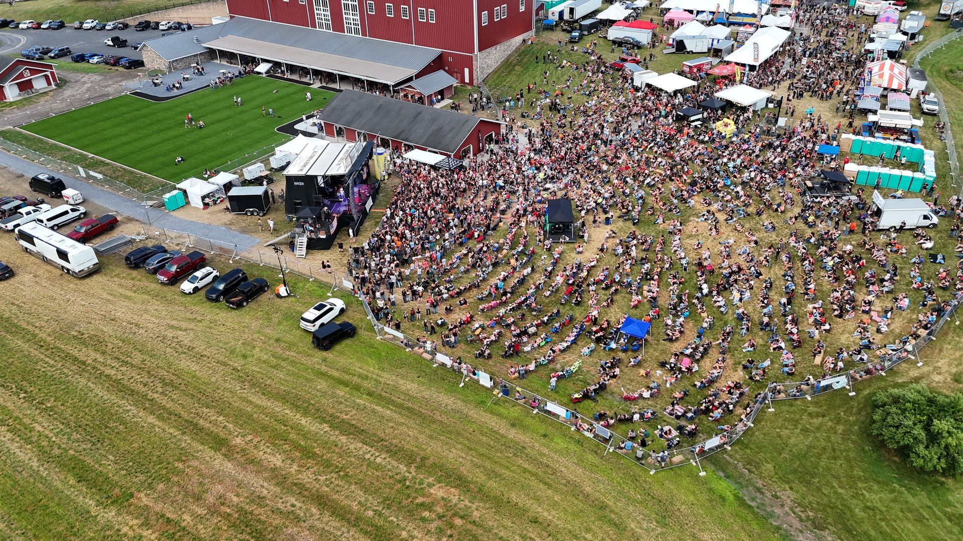Large outdoor concert; crowd gathered on a grassy field facing a stage near a red barn.