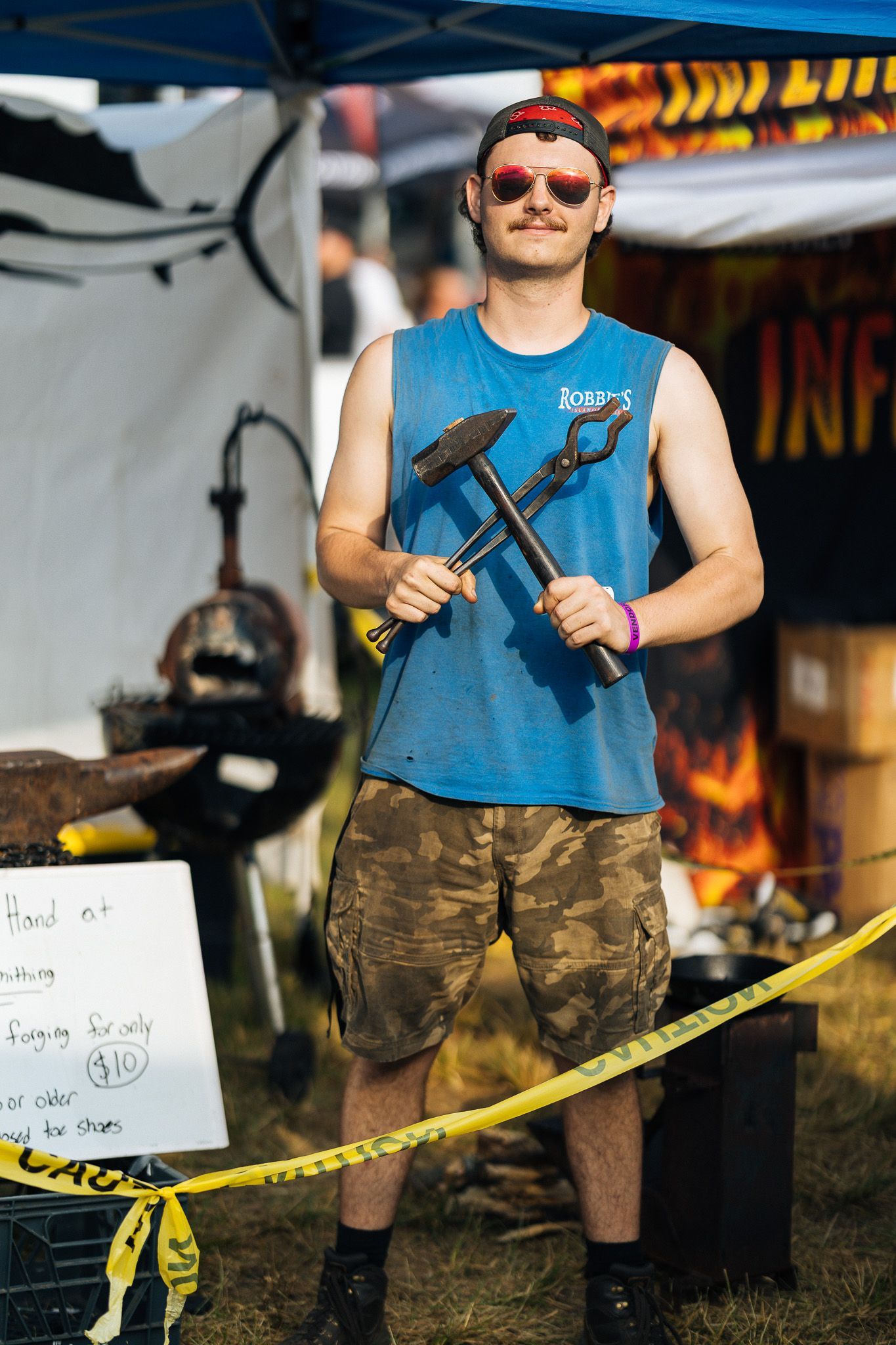 A blacksmith holding tools, wearing blue tank top, camo shorts, at a fair.