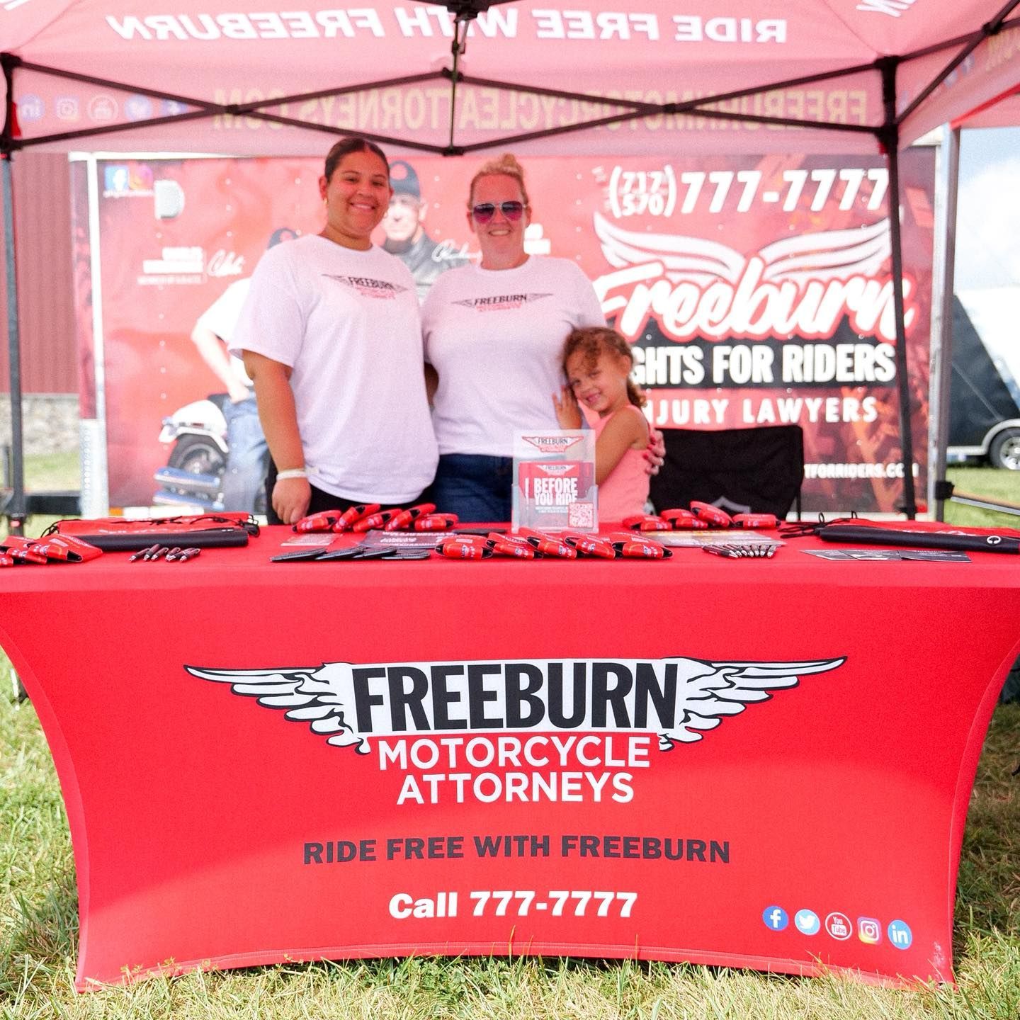 Three women at a red Freeburn Motorcycle Attorneys booth. One has a child. They smile. Red and white colors.