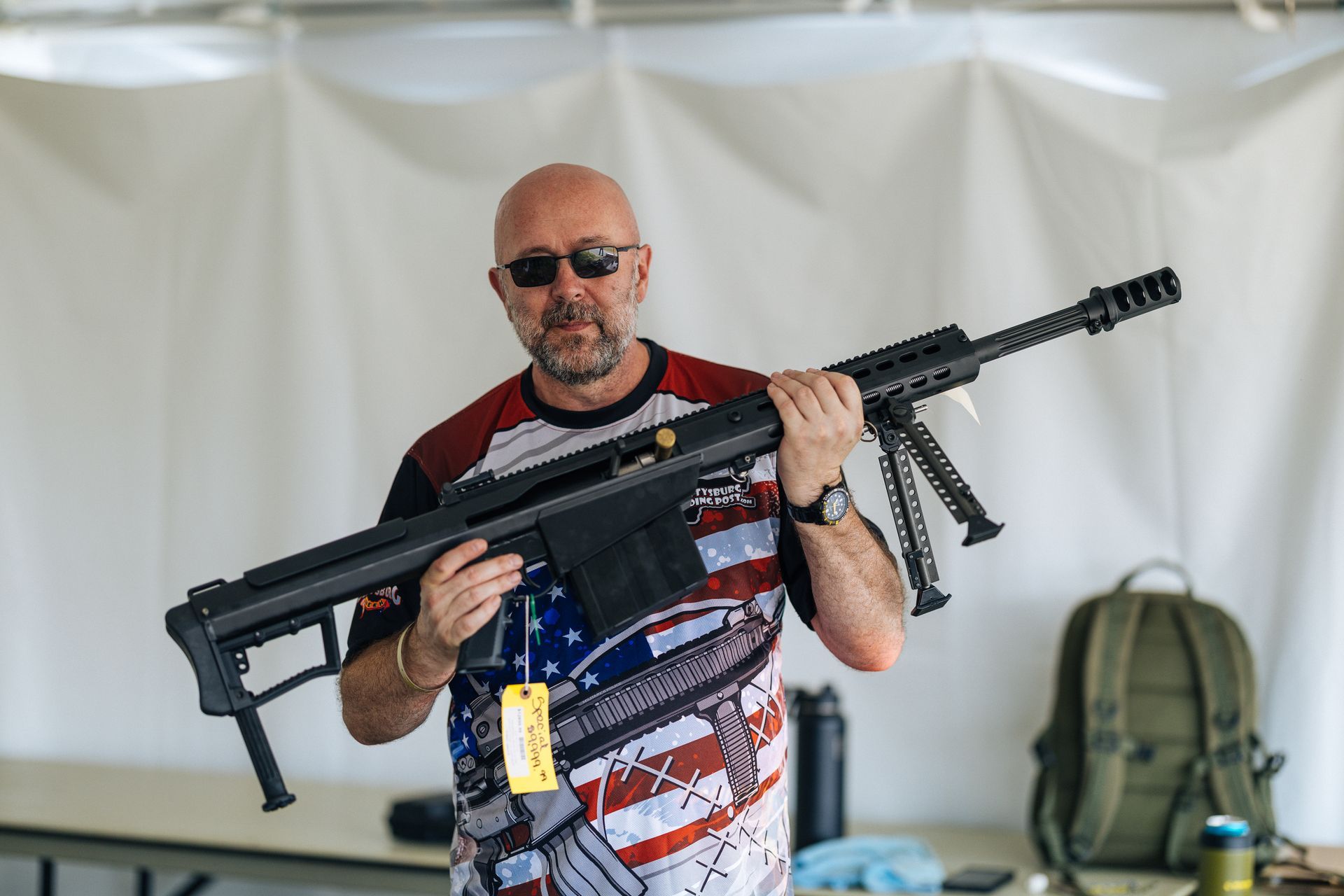 Man holding a large black rifle with bipod, wearing sunglasses and a patriotic shirt.