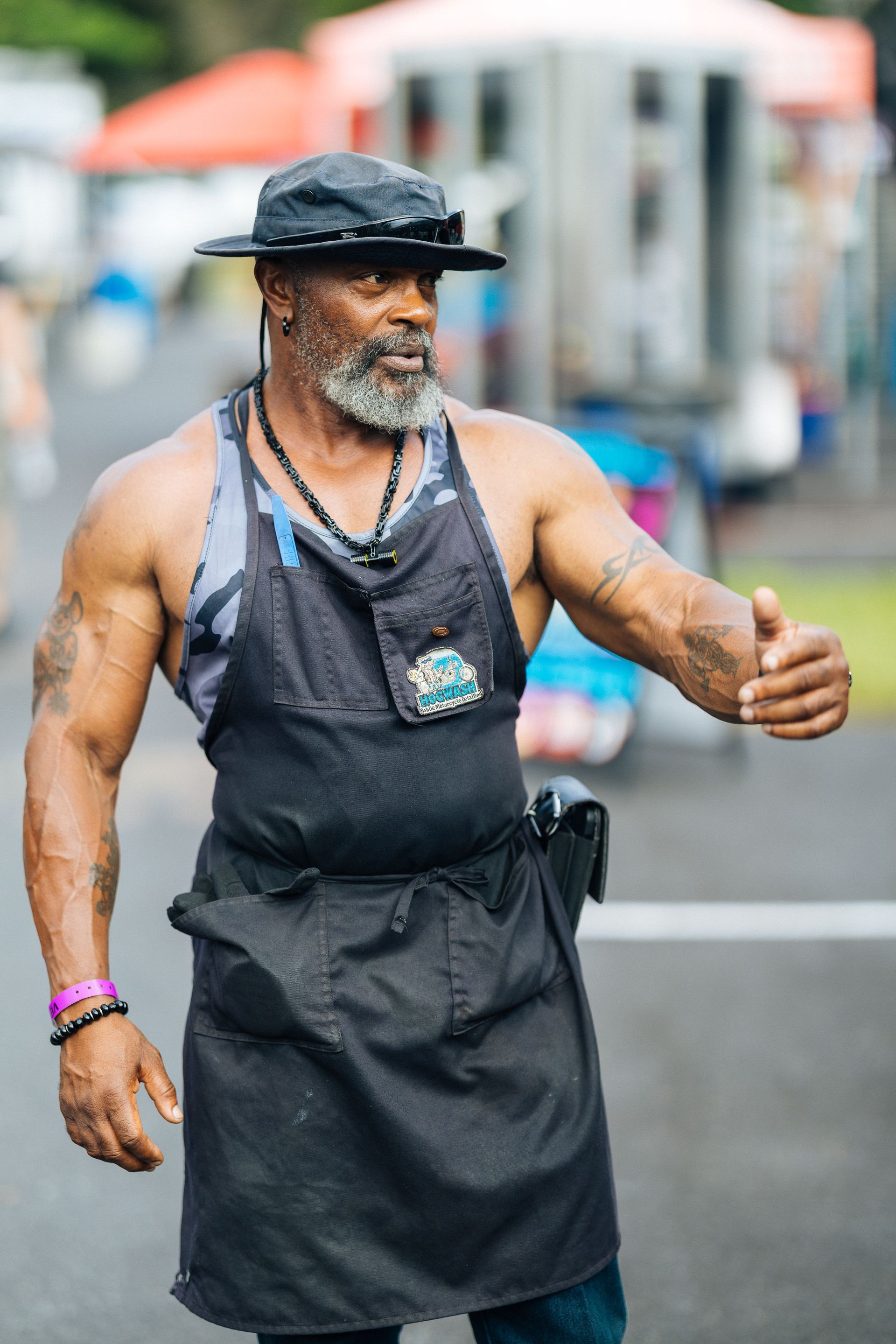 Man in apron and hat gestures with hand; outdoor market setting.