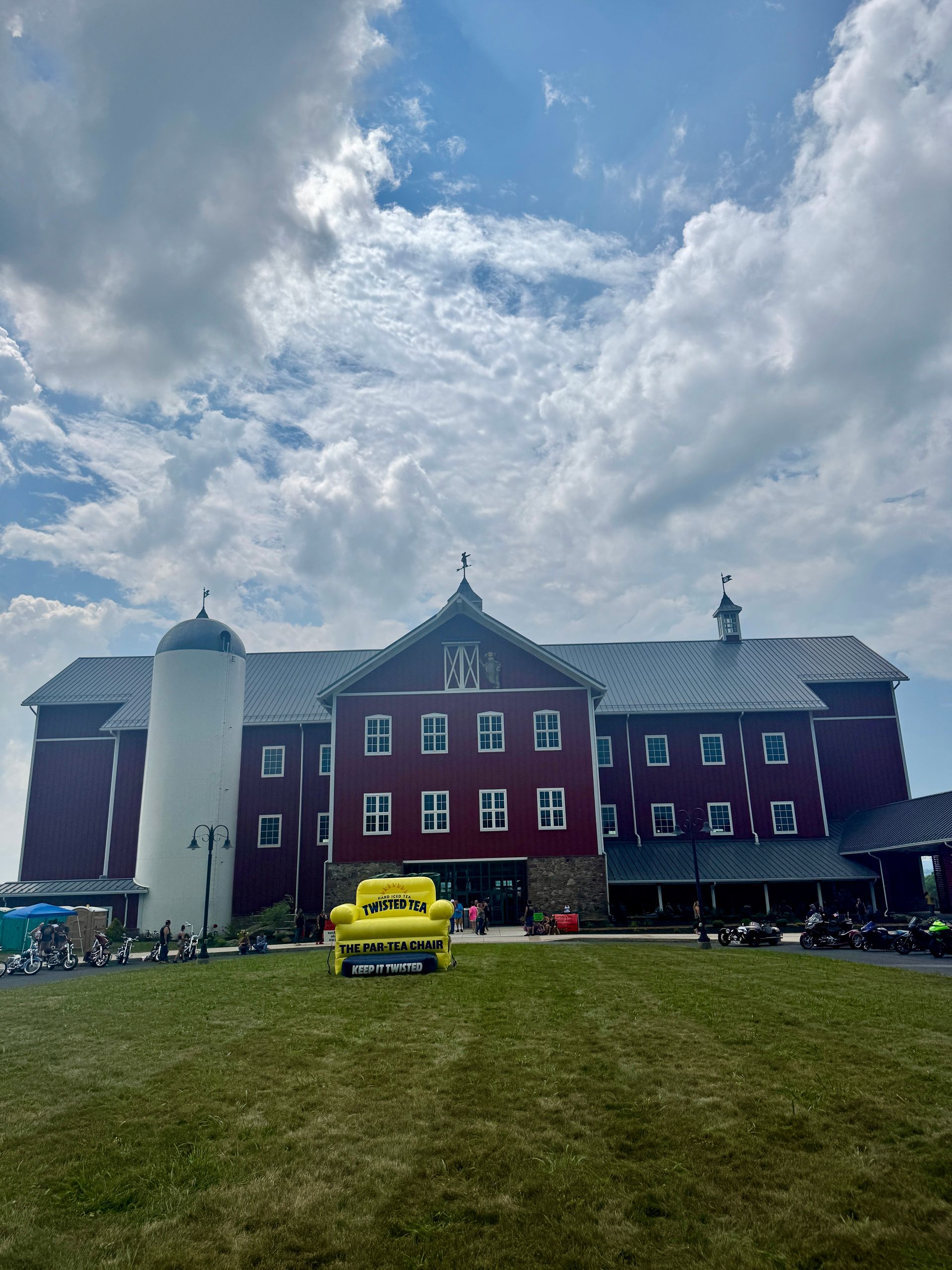 Red barn with silo, yellow truck on green lawn, cloudy sky.