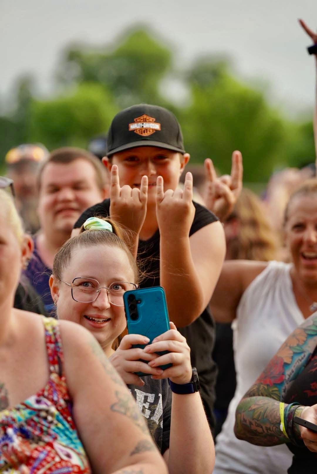 A crowd at a concert with a young person making a rock on gesture with their hands, a woman smiling and taking a photo.