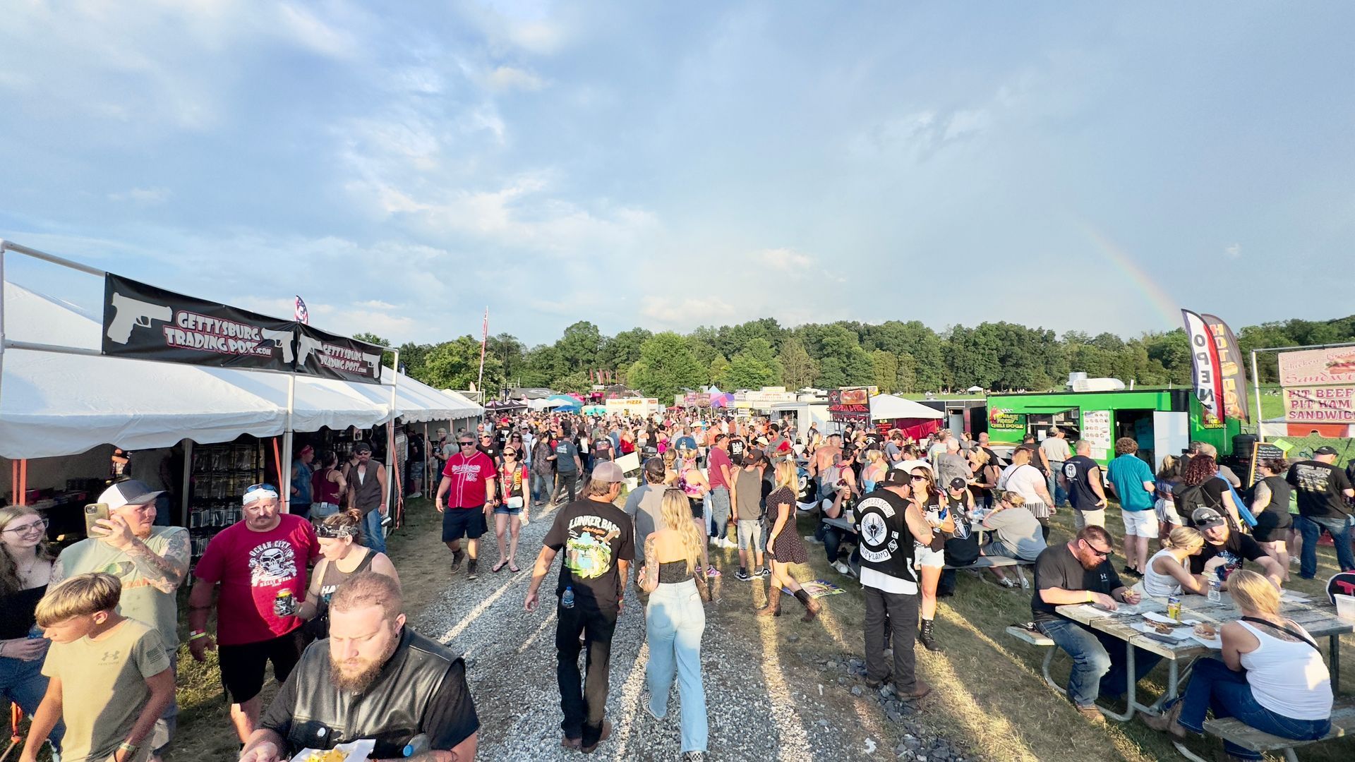 Crowd at an outdoor festival, tents, food vendors, people walking and socializing. Sky with rainbow.