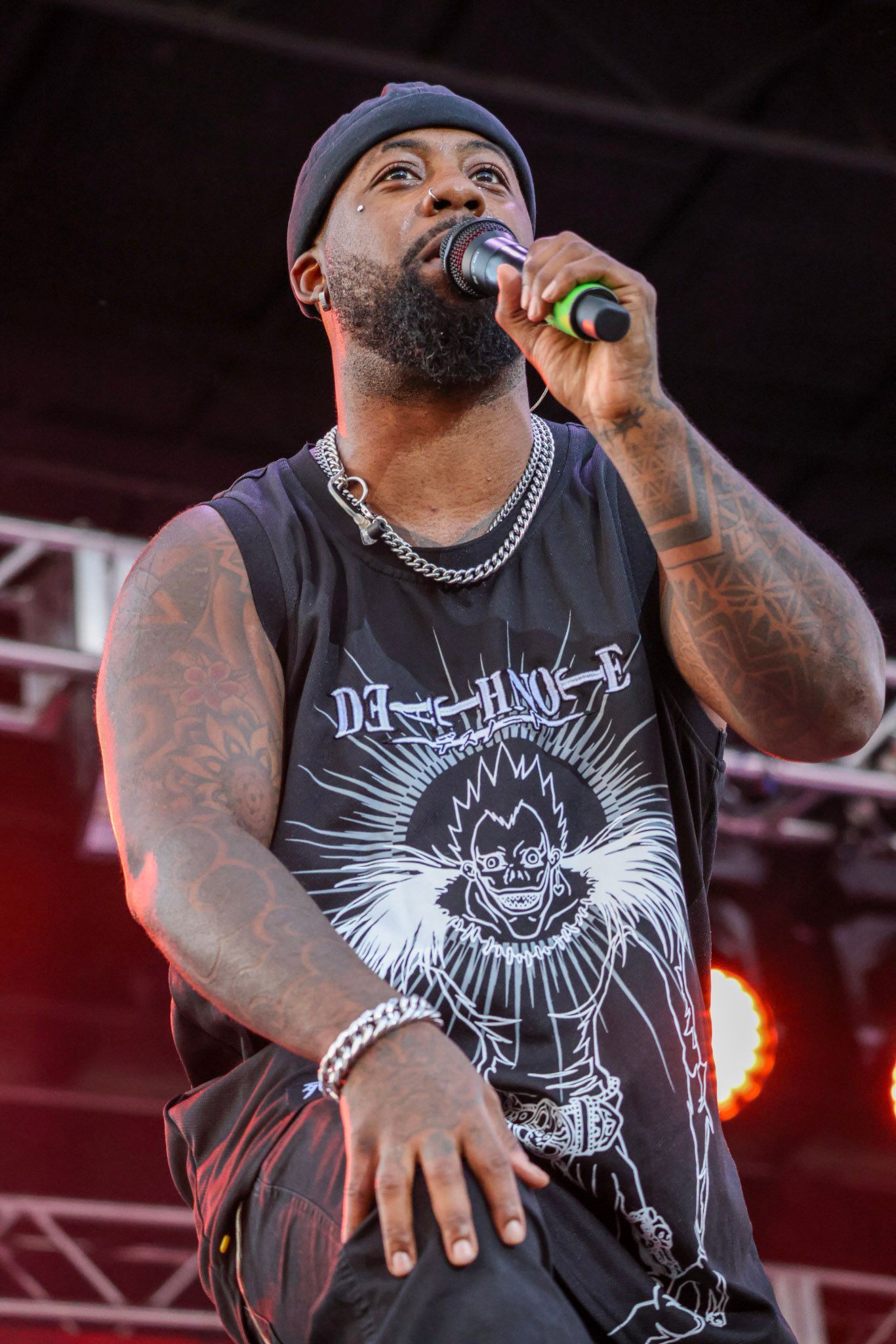 Man with beard singing into microphone, wearing a beanie, tank top, and chain.