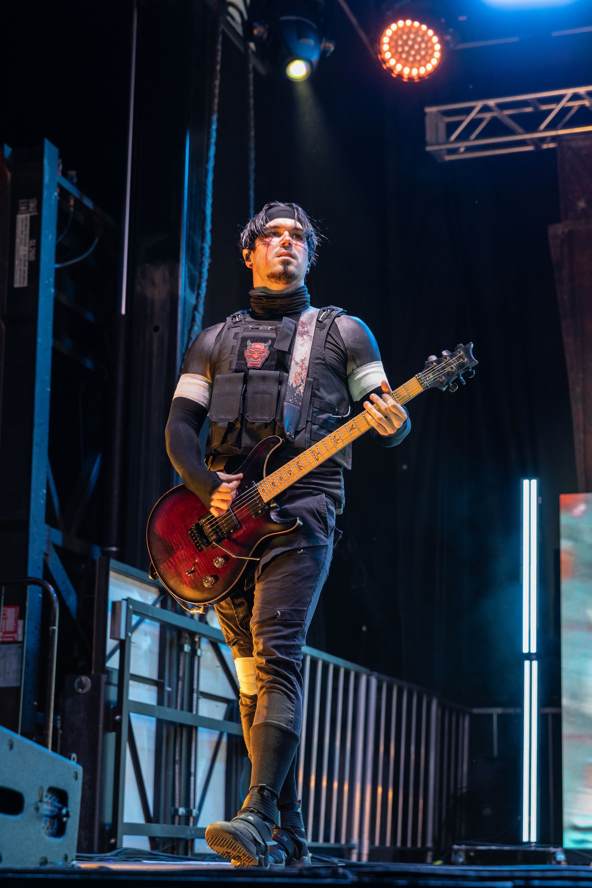 Guitarist on stage, wearing black tactical vest and wristbands, holding a red and black guitar.