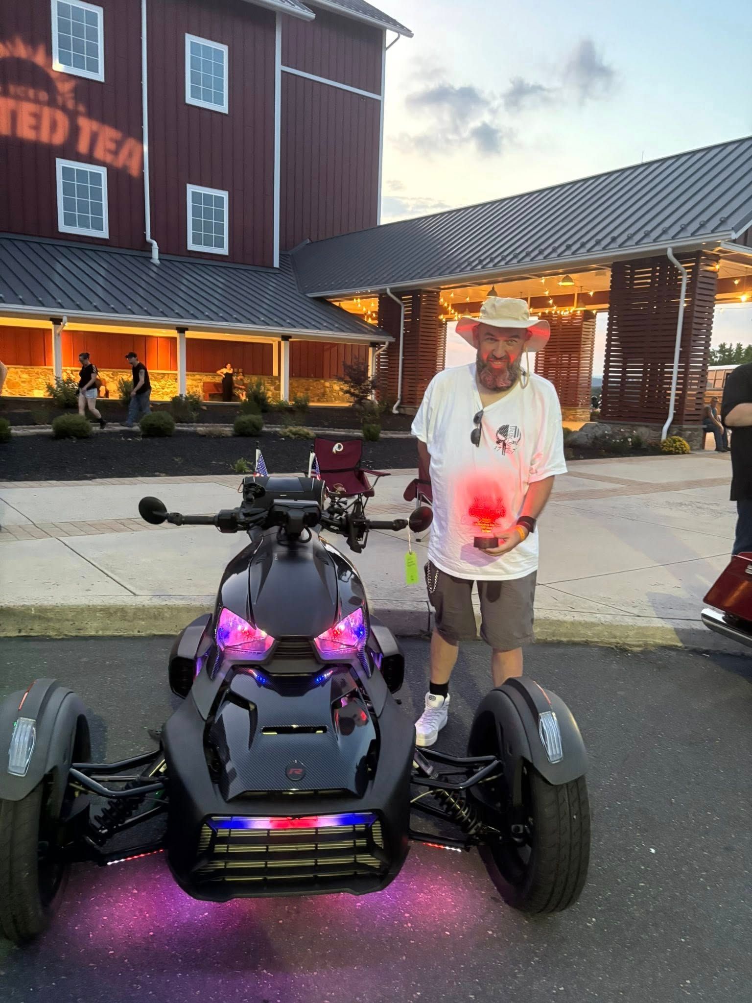 Man in hat stands by black three-wheeled motorcycle with colorful lights, red building background.