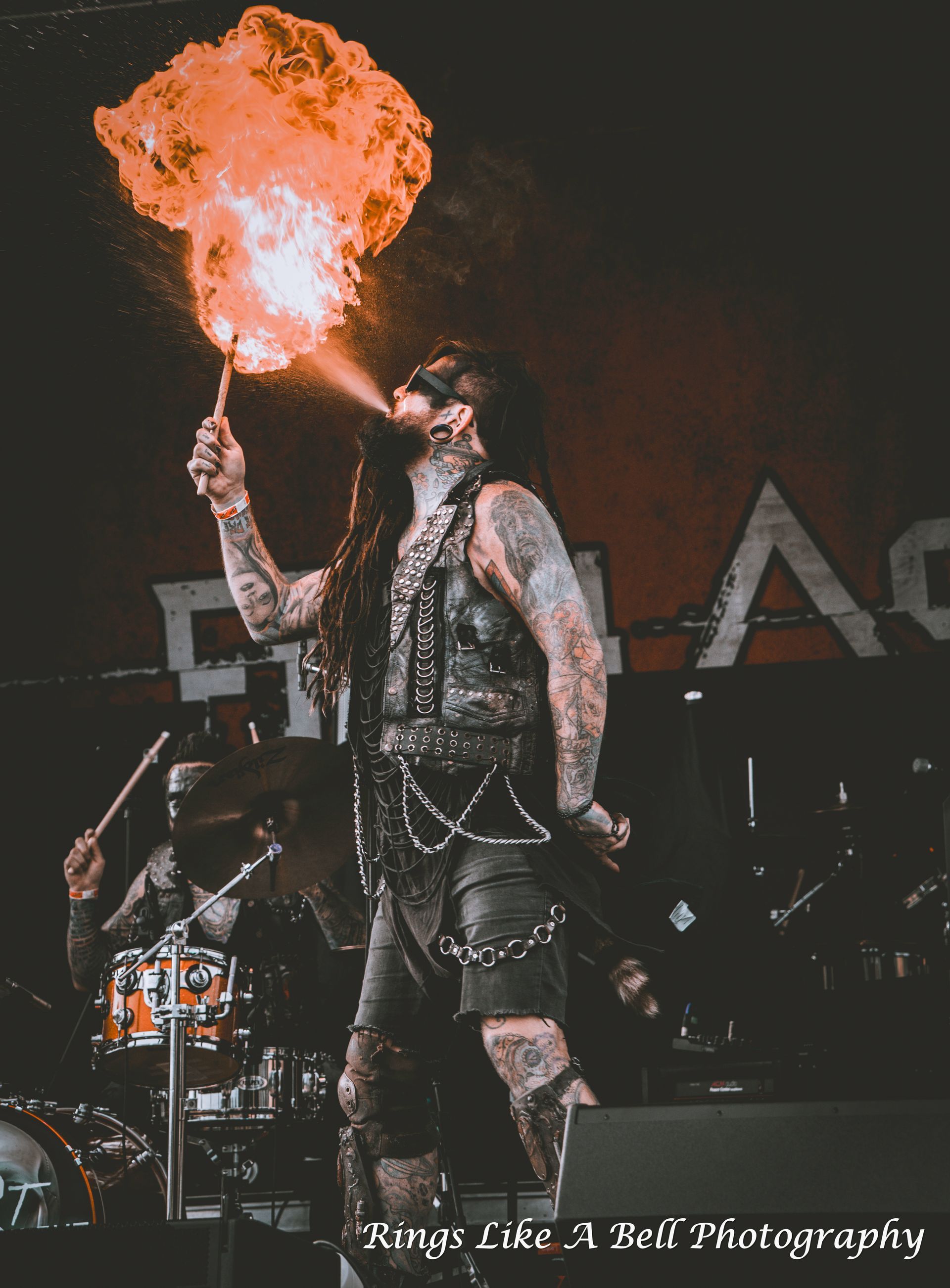 Man breathing fire on stage, tattoos, leather outfit. Drummer in background.