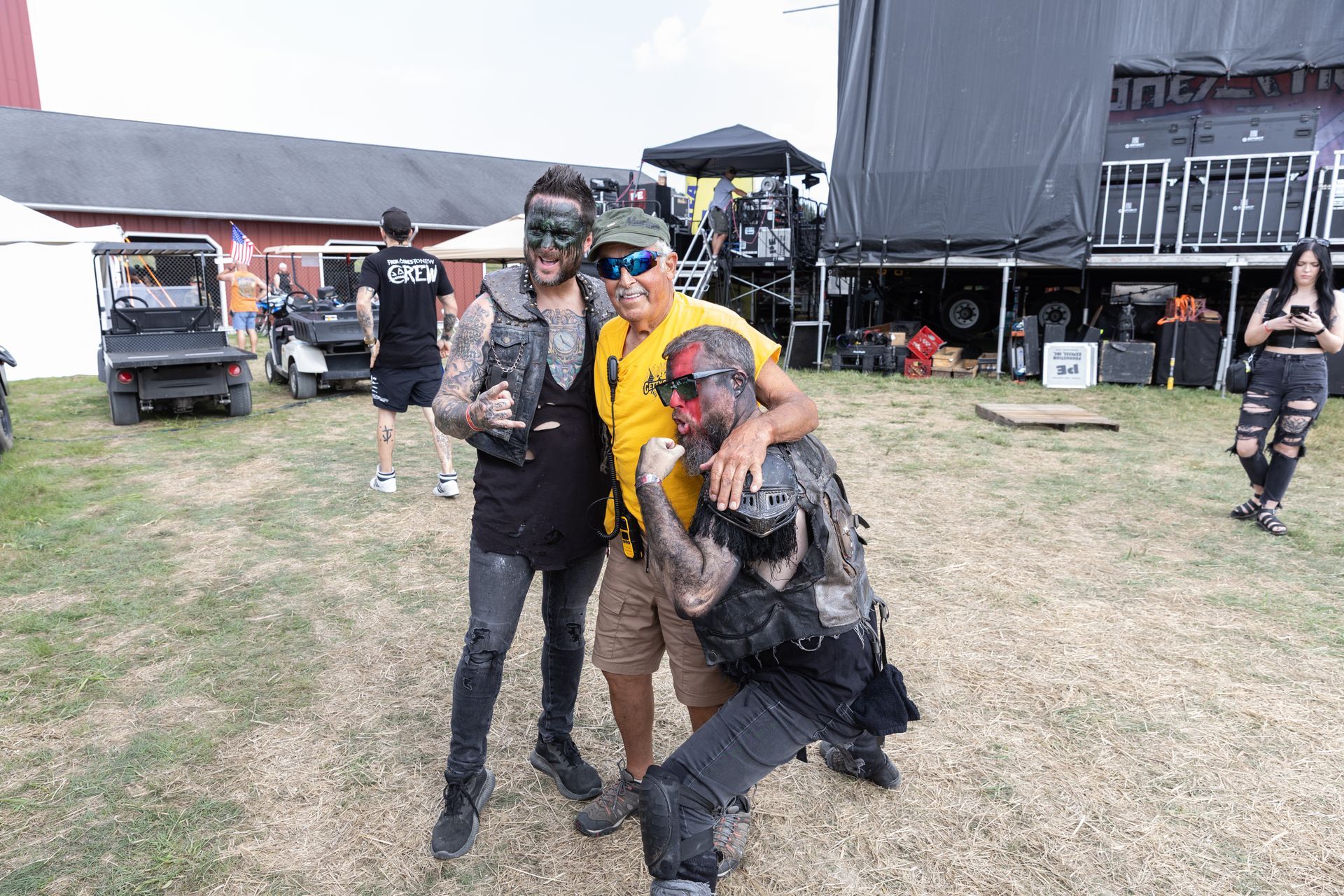 Three people pose for a photo outside. Two wear dark stage makeup and outfits. A man in a yellow shirt embraces them.