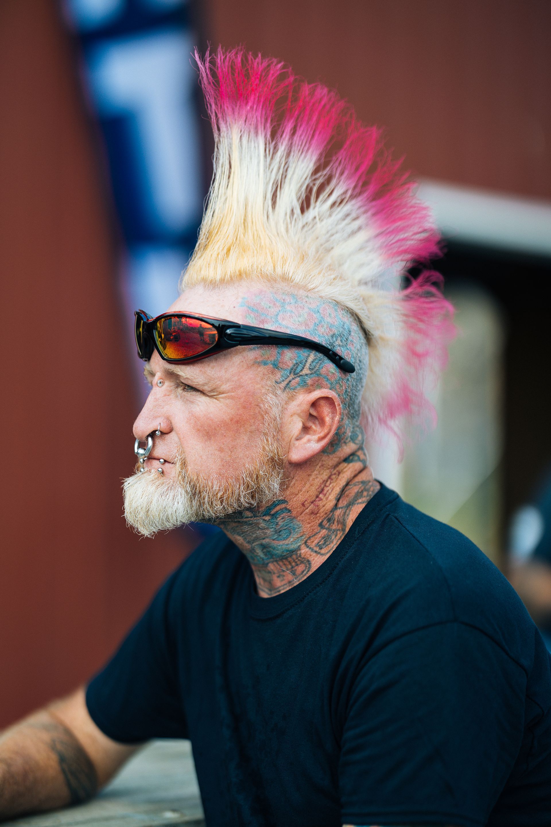 Man with a pink and yellow mohawk, tattoos, and piercings wearing sunglasses.