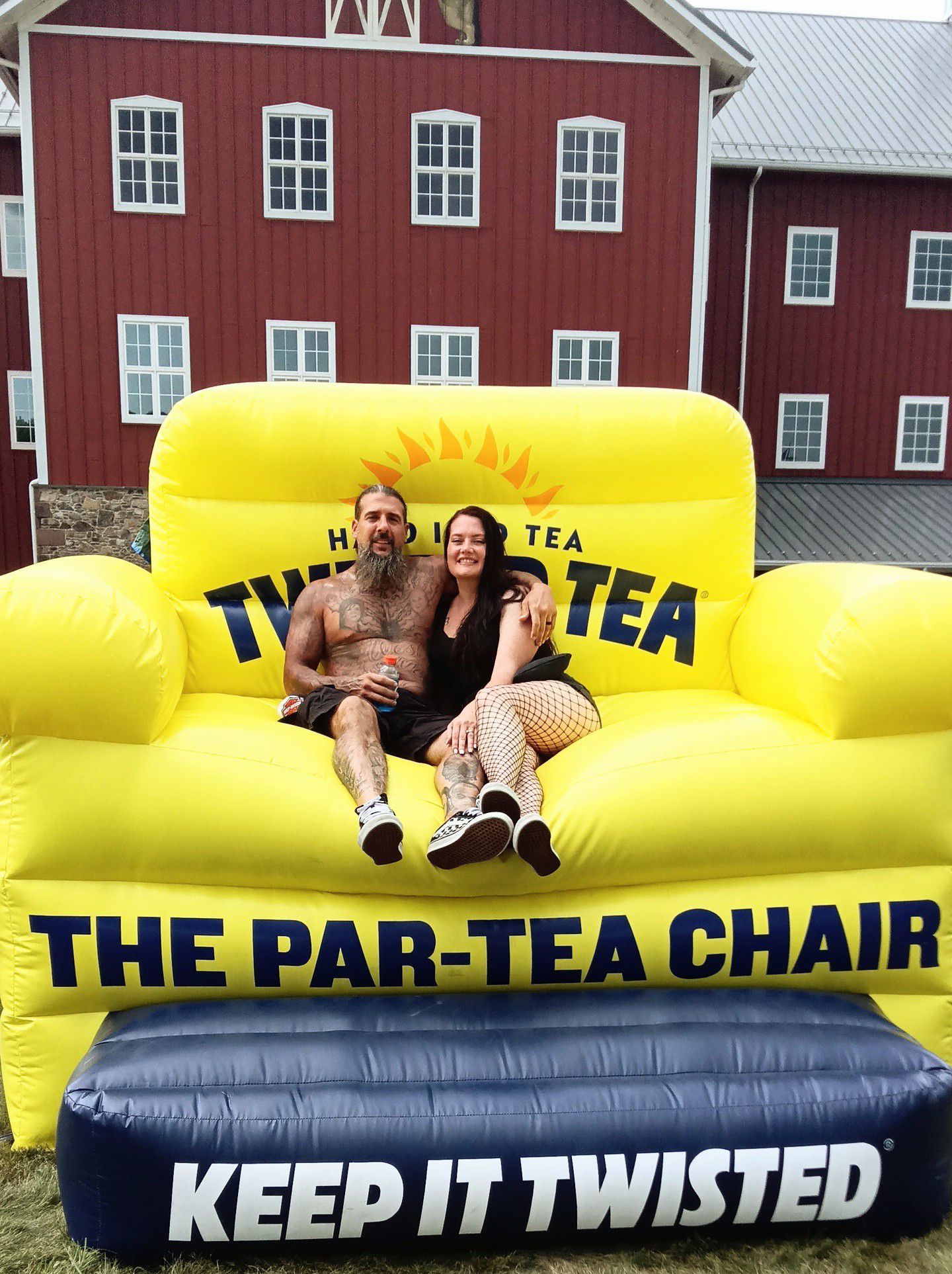 Two people on a large inflatable Twisted Tea chair in front of a red barn.