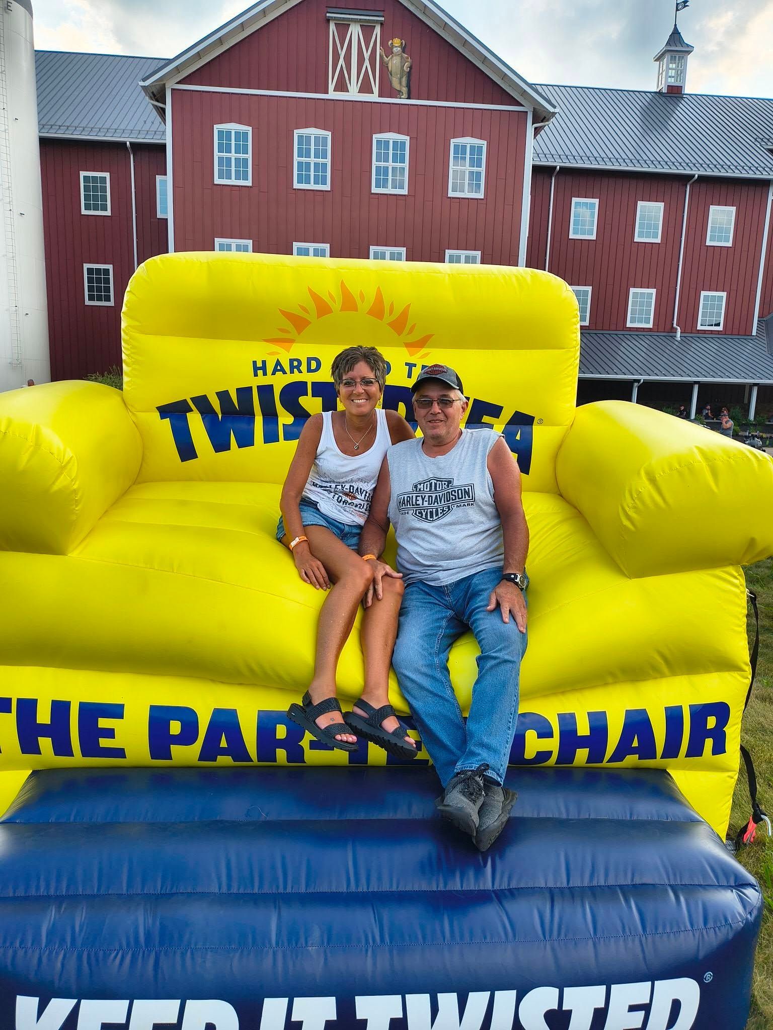 Two people sit on an inflatable Twisted Tea chair in front of a red barn.